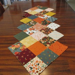 May include: A quilted table runner with a diamond pattern. The runner features squares of fabric in various colors, including orange, yellow, green, and burgundy, with floral and patterned designs. The runner is placed on a wooden table.