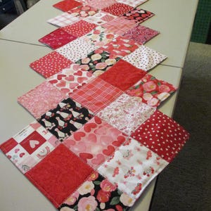 May include: A quilted table runner featuring a patchwork design with various red, pink, and white fabrics. Patterns include hearts, polka dots, floral prints, and plaid. The runner is displayed on a light-colored table.