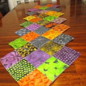 May include: A colorful Halloween-themed table runner. The runner is made of quilted fabric squares in orange, purple, green, and gray. The squares feature patterns of bats, spiders, webs, and polka dots. The runner is displayed on a dark wooden table.