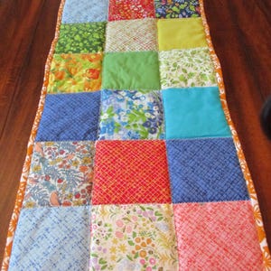May include: A colorful patchwork table runner with a variety of patterned squares in shades of orange, blue, green, red, and yellow. The runner has an orange scalloped border and the date 07/12/2025 is visible in the bottom right corner.