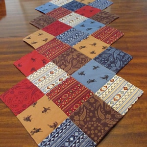 May include: A patchwork table runner with a diamond pattern. The squares feature various designs, including paisley, geometric patterns, and silhouettes of horses and riders. The color palette includes red, blue, brown, and tan.