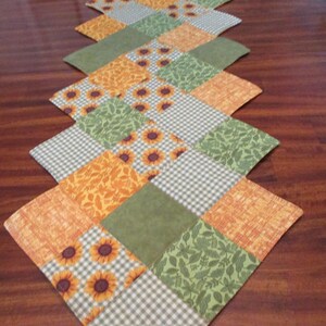 May include: A quilted table runner with a diamond pattern. The runner features squares of orange, green, and white fabric. Some squares have sunflower, leaf, and gingham patterns. The date 07/12/2025 is printed on the bottom right.
