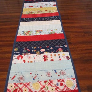 May include: A patriotic table runner with a patchwork design. The runner features red, white, and blue fabrics with patterns of fireworks, watermelon, and floral prints. The runner is approximately 12 inches wide and 48 inches long.