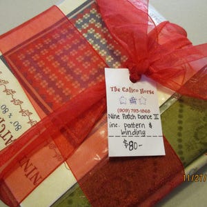 May include: A fabric bundle wrapped with a red ribbon. The bundle includes patterned fabric squares in red, green, and blue hues. A tag reads "Nine Patch Dance II" and includes pattern and binding.