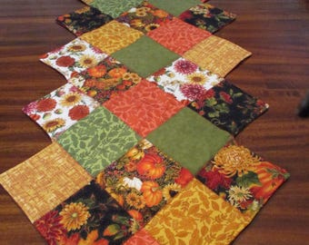 May include: A quilted table runner featuring a patchwork design with various autumn-themed fabrics. The runner showcases squares of orange, green, yellow, and black fabrics with floral, leaf, and pumpkin patterns. The date "10/30/2024" is visible.
