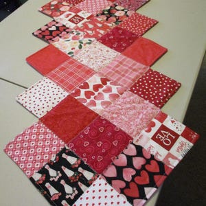 May include: A red and pink patchwork table runner with a variety of heart patterns. The runner is made of different fabrics, including plaid, polka dots, and floral prints. The runner is approximately 18 inches wide and 60 inches long.