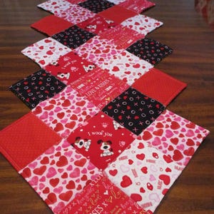 May include: A quilted table runner with a patchwork design. The runner features red, white, and black squares with heart and dog patterns, and text that says "Love & Kisses" and "I woof you".
