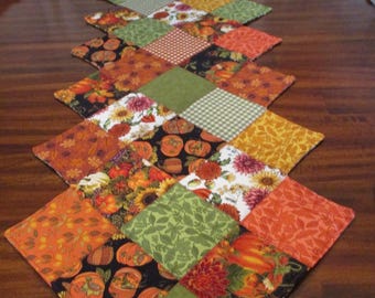 May include: A quilted table runner featuring a diamond pattern. The runner showcases a variety of autumn-themed fabrics, including pumpkins, sunflowers, and fall leaves in shades of orange, green, and yellow. The date "10/30/2024" is visible.
