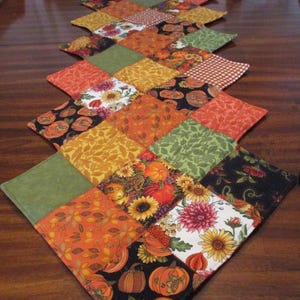May include: A quilted table runner with a diamond pattern. The runner features squares of fabric in autumn colors, including orange, green, yellow, and black. Some squares have floral and pumpkin patterns. The date "10/30/2024" is visible.