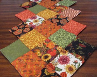 May include: A quilted table runner with a diamond pattern. The runner features squares of fabric in autumn colors, including orange, green, yellow, and black. Some squares have floral and pumpkin patterns. The date "10/30/2024" is visible.
