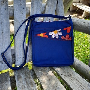 Puede incluir: Un bolso bandolera de lona azul con un diseño floral rojo, blanco y amarillo. El bolso tiene una correa larga ajustable.