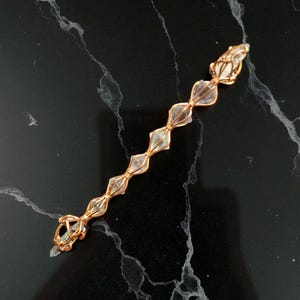 May include: A linear jewelry piece featuring seven clear, faceted crystal beads secured in a row by copper-colored wire wrapping. The wire forms a decorative cage around each individual crystal, connecting them in a continuous, elongated chain design.