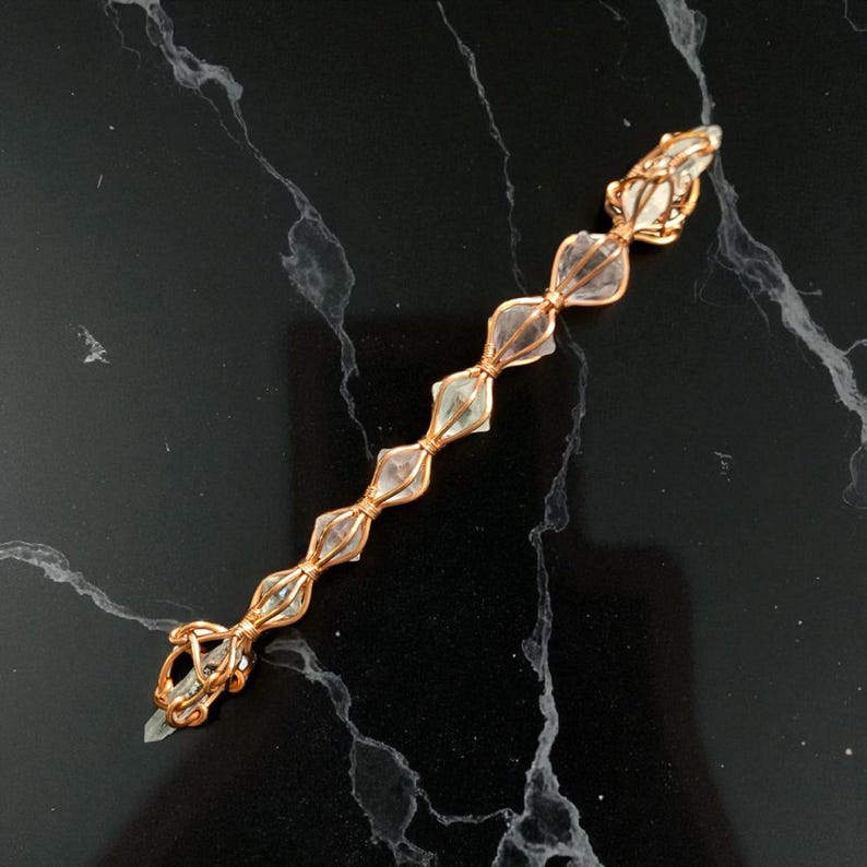 May include: A linear jewelry piece featuring seven clear, faceted crystal beads secured in a row by copper-colored wire wrapping. The wire forms a decorative cage around each individual crystal, connecting them in a continuous, elongated chain design.