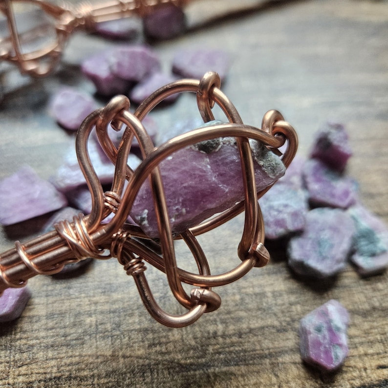 May include: A raw, purplish-pink crystal stone encased in a decorative cage made of coiled copper wire. The wire is wrapped tightly around the base of the crystal, forming a series of loops that secure the stone within the metal structure.