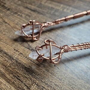 May include: Two hair sticks featuring clear, pointed quartz crystals secured in intricate, hand-wrapped copper wire cages. The copper wire extends down the length of each stick, showcasing a detailed, coiled metal design that holds the stones.