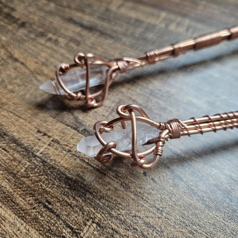 May include: Two hair sticks featuring clear, pointed quartz crystals secured in intricate, hand-wrapped copper wire cages. The copper wire extends down the length of each stick, showcasing a detailed, coiled metal design that holds the stones.