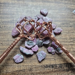 May include: Two copper wire-wrapped wands featuring raw, pinkish-purple gemstone clusters at the tips. The metal handles are tightly coiled with copper wire, and the wands are displayed alongside several loose, unpolished pink and grey stone fragments.