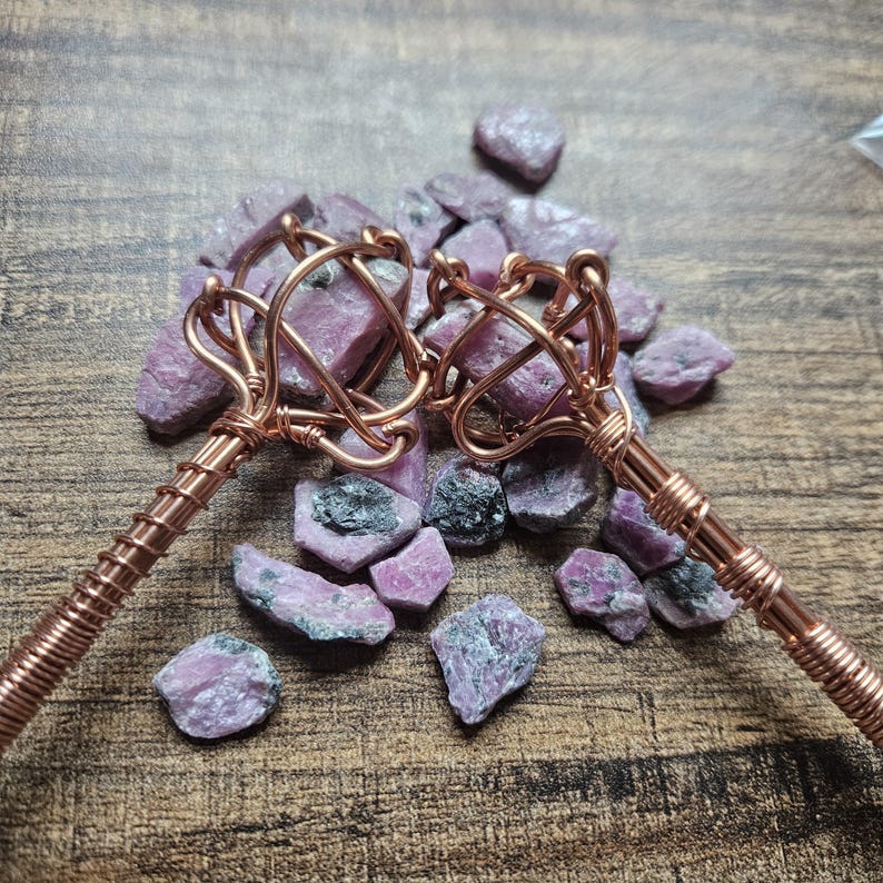 May include: Two copper wire-wrapped wands featuring raw, pinkish-purple gemstone clusters at the tips. The metal handles are tightly coiled with copper wire, and the wands are displayed alongside several loose, unpolished pink and grey stone fragments.