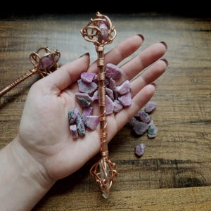 May include: A copper wire-wrapped wand featuring a raw pink and grey stone at the top and a clear crystal point at the base. The handle is constructed from multiple strands of coiled copper wire, surrounded by several loose, raw pink stone fragments.