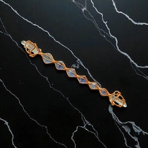 May include: A long, slender wand-like object featuring a clear, pointed crystal encased in a decorative copper wire wrap. The wire forms a series of repeating diamond-shaped loops along the length of the crystal, securing several smaller, translucent, light-blue, faceted stones within the metallic structure.