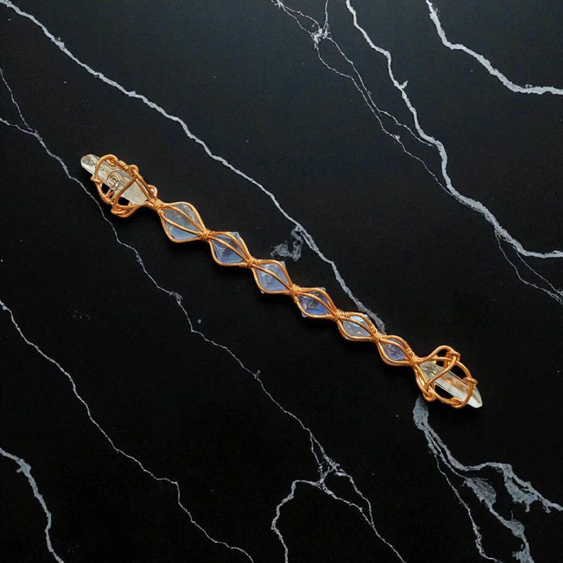 May include: A long, slender wand-like object featuring a clear, pointed crystal encased in a decorative copper wire wrap. The wire forms a series of repeating diamond-shaped loops along the length of the crystal, securing several smaller, translucent, light-blue, faceted stones within the metallic structure.