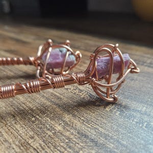 May include: Two hair sticks crafted from copper wire wrapped around raw, pink-colored crystals. The metal wire is coiled tightly along the length of the sticks and forms an intricate, cage-like structure around each of the rough, rectangular stones.