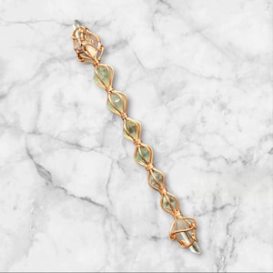 May include: A long, slender wand-like object featuring a series of six pale green, translucent, faceted crystals encased in a decorative, intricate copper wire wrap that forms a repeating cage pattern along the entire length of the piece.