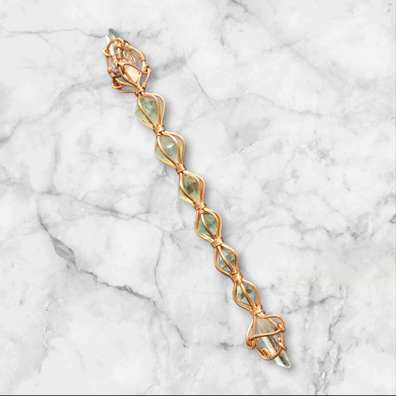 May include: A long, slender wand-like object featuring a series of six pale green, translucent, faceted crystals encased in a decorative, intricate copper wire wrap that forms a repeating cage pattern along the entire length of the piece.