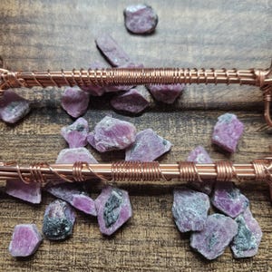 May include: Two parallel copper wire structures featuring intricate wire-wrapped detailing, positioned over a scattered collection of raw, unpolished pink and grey ruby gemstone fragments with visible crystalline textures and natural inclusions.