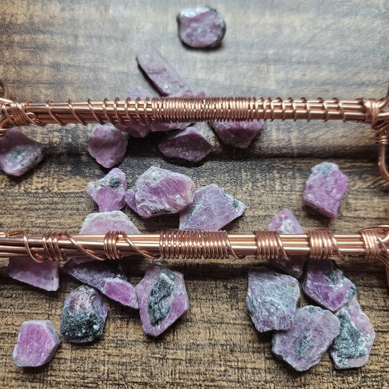 May include: Two parallel copper wire structures featuring intricate wire-wrapped detailing, positioned over a scattered collection of raw, unpolished pink and grey ruby gemstone fragments with visible crystalline textures and natural inclusions.