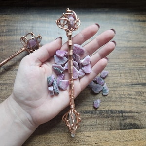 Op de afbeelding: Een met koperdraad gewikkelde toverstaf met een heldere kristallen punt en een roze kristal aan de bovenkant. De toverstaf is omgeven door kleine roze en groene kristallen.