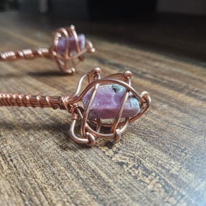 May include: Two hair sticks featuring raw, pink, rectangular-shaped crystals encased in intricate, spiraled copper wire cages. The copper wire is tightly wrapped around the stems of the sticks, providing a metallic, reddish-brown finish.