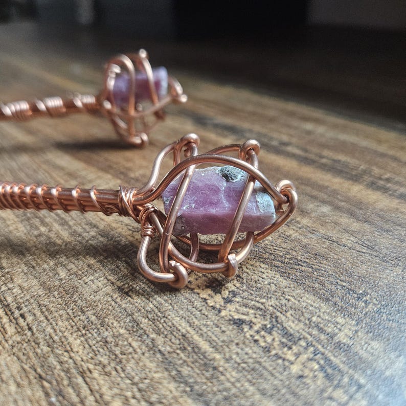 May include: Two hair sticks featuring raw, pink, rectangular-shaped crystals encased in intricate, spiraled copper wire cages. The copper wire is tightly wrapped around the stems of the sticks, providing a metallic, reddish-brown finish.