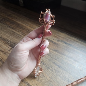 May include: A handcrafted wand featuring a central copper wire shaft wrapped in intricate metal coils. The top is adorned with a pink and grey stone encased in a wire cage, while the bottom features a clear, pointed crystal held by copper wire.