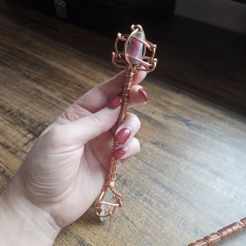 May include: A handcrafted wand featuring a central copper wire shaft wrapped in intricate metal coils. The top is adorned with a pink and grey stone encased in a wire cage, while the bottom features a clear, pointed crystal held by copper wire.