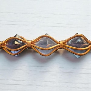 May include: A horizontal jewelry piece featuring three faceted, translucent purple crystals encased in a decorative copper wire wrap. The wire is coiled tightly between each crystal, creating a repeating oval pattern that secures the stones.