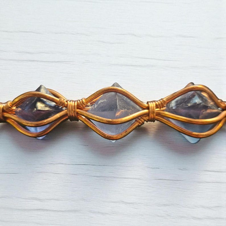 May include: A horizontal jewelry piece featuring three faceted, translucent purple crystals encased in a decorative copper wire wrap. The wire is coiled tightly between each crystal, creating a repeating oval pattern that secures the stones.