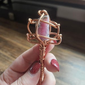 May include: A raw pink crystal point encased in a decorative copper wire wrap. The metal wire is coiled around the stone and extends down into a straight, tightly wound copper handle, creating a wand-like structure with a metallic finish.