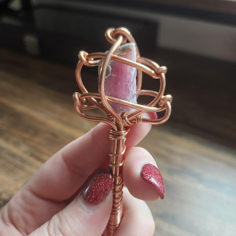May include: A raw pink crystal point encased in a decorative copper wire wrap. The metal wire is coiled around the stone and extends down into a straight, tightly wound copper handle, creating a wand-like structure with a metallic finish.