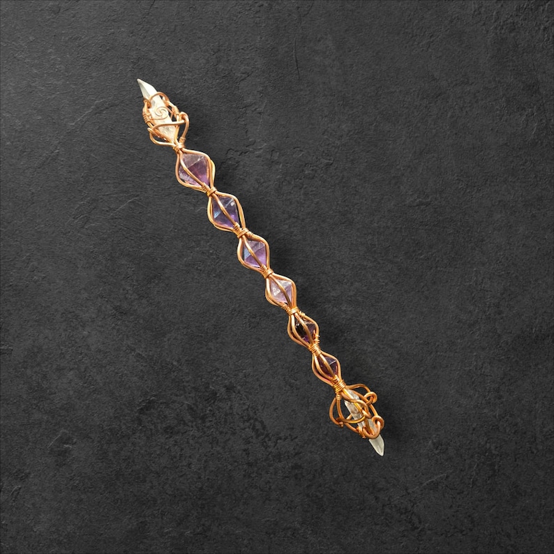 May include: A long, slender wand-like object featuring a central core of clear, pointed crystals encased in a decorative, spiraling copper wire cage. The wire framework holds several small, faceted, light purple amethyst stones in a linear row.