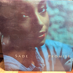 May include: Album cover for Sade's "Promise." The cover features a close-up of a woman's face with a soft, diffused aesthetic. The title "Sade" and the album title "Promise" are printed in a simple, elegant font.