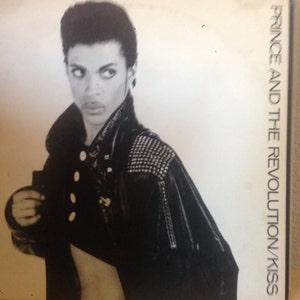 May include: Black and white photo of Prince and The Revolution on a vinyl record cover. The cover features a close-up of Prince wearing a black leather jacket with studs. The text "Prince and The Revolution/Kiss" is printed on the right side of the cover.