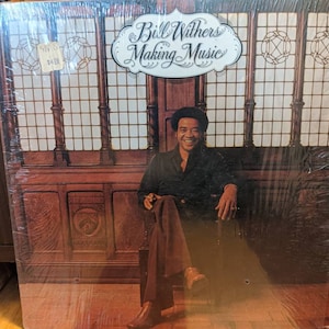 May include: A vinyl record album cover titled Bill Withers Making Music. The cover features a smiling person in a dark shirt and brown trousers sitting in a chair against a background of dark wood panelling and a grid-patterned window.