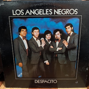 Peut inclure: Une pochette de disque vinyle vintage représentant le groupe Los Angeles Negros. La pochette montre cinq personnes en costumes rayés sombres assortis avec chemises blanches et nœuds papillon sur un fond bleu nuageux. Le texte indique Los Angeles Negros et Despacito.