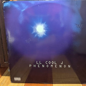 May include: Album cover for LL Cool J's "Phenomenon." The cover features a dark blue and purple gradient background with a bright white light in the center. The album title and artist name are in white text.