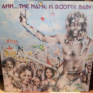 May include: A vintage vinyl record album cover featuring a colorful illustration of a man with a large afro wearing a white shirt and a beaded necklace. The album title is "Ahh...The Name Is Bootsy, Baby!" and the background is a vibrant mix of colors and shapes.