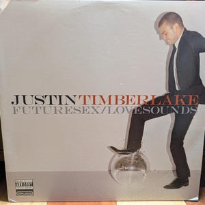 May include: Album cover for Justin Timberlake's "FutureSex/LoveSounds." The image features Timberlake in a black suit, stepping on a disco ball. The album title and artist name are printed in a mix of red and gray fonts against a light gray background.