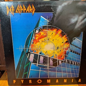 Könnte beinhalten: Ein Vintage-Vinyl-Plattencover mit der Band Def Leppard. Das Cover zeigt eine blaue und gelbe Illustration eines Wolkenkratzers mit einem Ziel und Flammen. Der Albumtitel "Pyromania" ist in großen weißen Buchstaben am unteren Rand des Covers gedruckt.