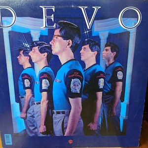 Op de afbeelding: Albumhoes van Devo, getiteld "New Traditionalists". De afbeelding toont vijf mannen in blauwe shirts en grijze broeken, in een rij. De bandnaam "DEVO" staat in het wit bovenaan.