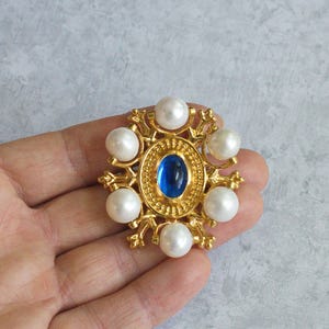 Puede incluir: Broche dorado con una gema azul ovalada central, rodeada de perlas blancas de imitación y detalles dorados ornamentados. El broche tiene un diseño clásico y elegante, adecuado para ocasiones formales o para añadir un toque vintage a un atuendo.